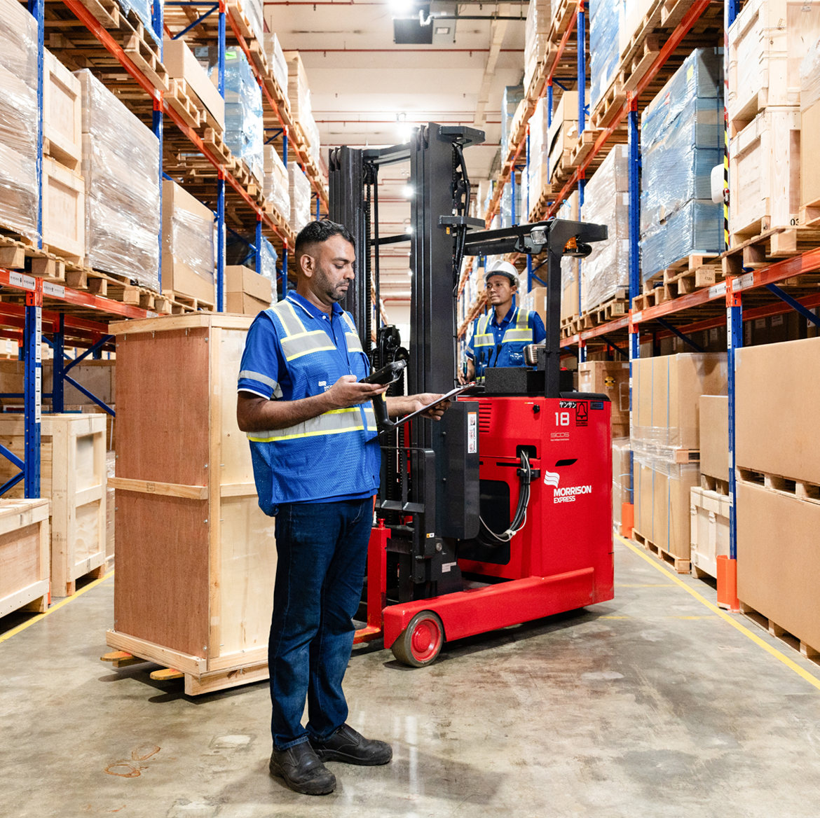 Two Morrison warehouse employees working in the warehouse, one driving a forklift unloading cargo pallets; the other scanning documents.