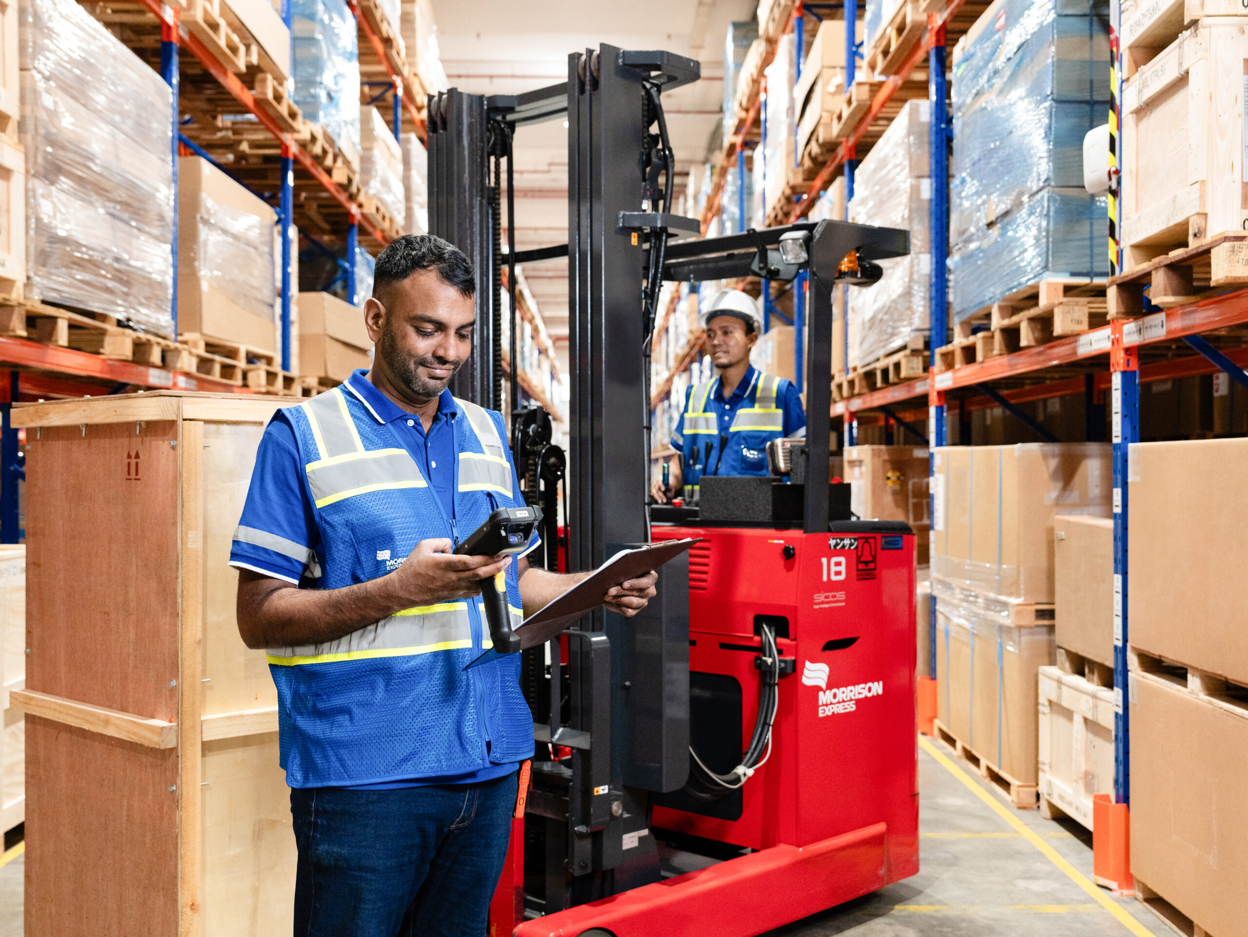 Two Morrison warehouse employees working in the warehouse, one driving a forklift unloading cargo pallets; the other scanning documents.