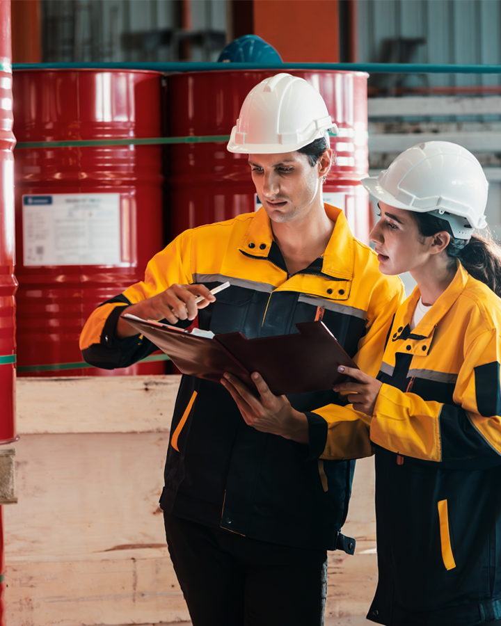 Two compliance officers by barrels of hazardous chemicals checking regulatory paperwork.