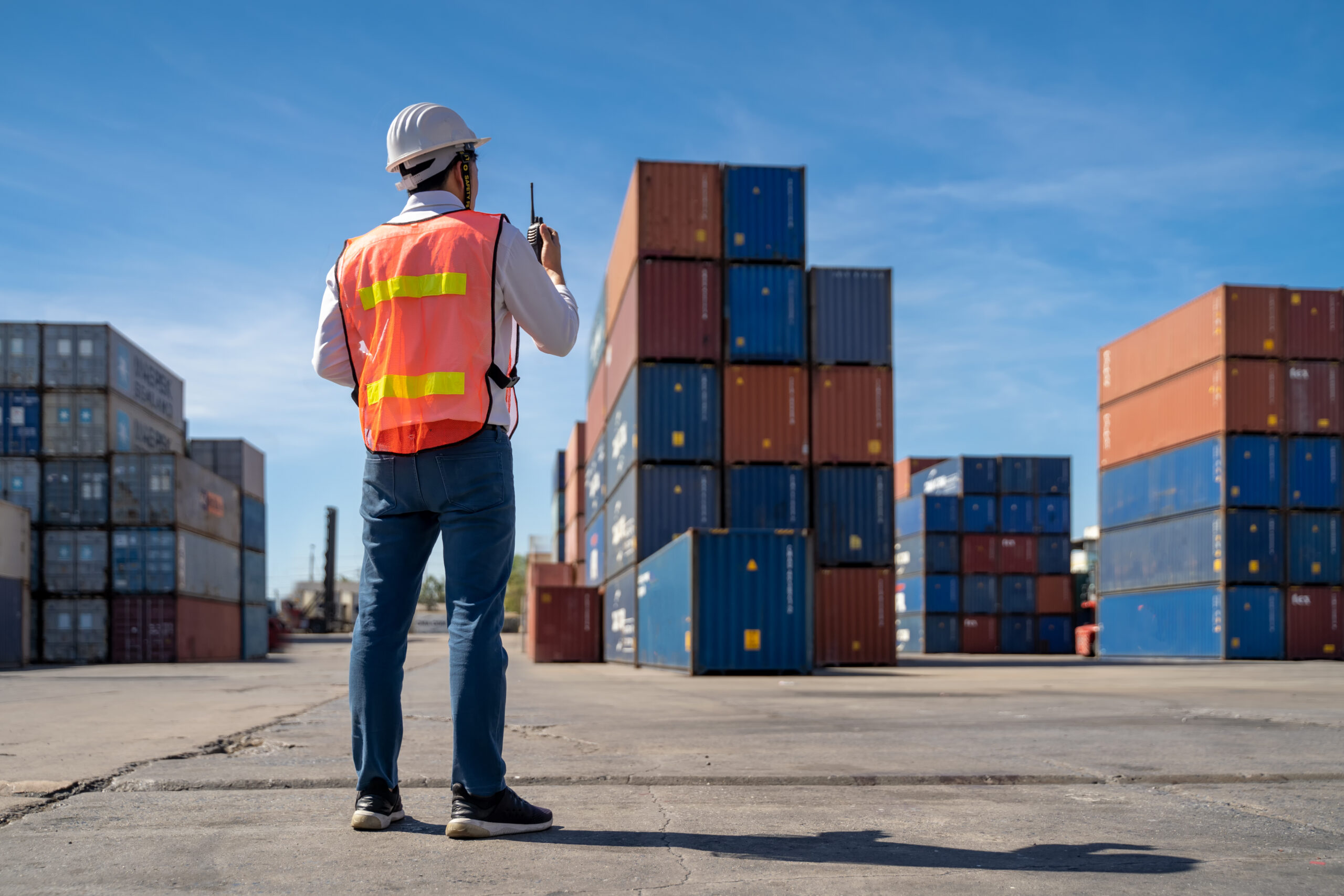 A logistics worker standing in a  container shipping yard communicating on a walkie-talkie. 