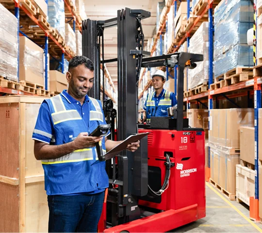 Two Morrison warehouse employees working in the warehouse, one driving a forklift unloading cargo pallets; the other scanning documents.