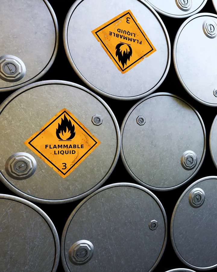 A stack of metal barrels containing chemicals labeled with orange "flammable liquid" labels.